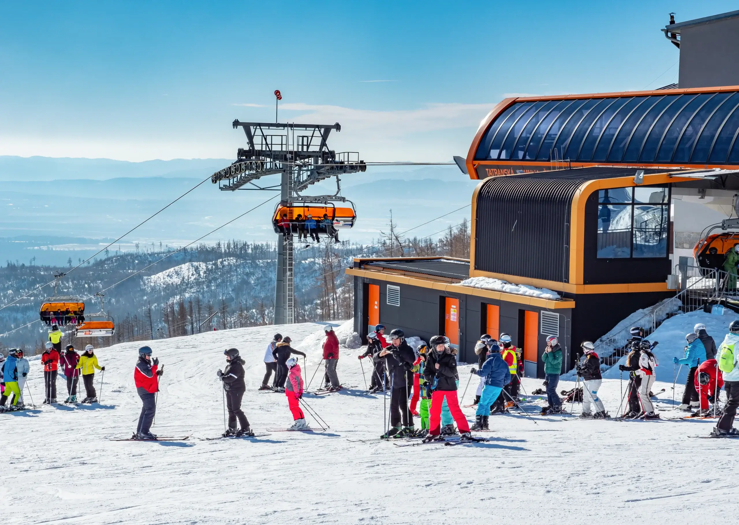 Et vinterlandskab med snedækkede bjerge i baggrunden, et skilt med teksten "Vysoké Tatry — Hory zážitkov" og "Tatranská Lomnica" samt en rød traktor af mærket Tatra i forgrunden.